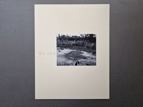 A black and white art print mounted on a wide off-white paper sheet. The print features a monochrome photograph of an outdoor scene with grass and puddles, with the handwritten text 'SON OF A BI'