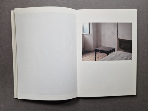 An open book or art print laid flat on a dark gray surface, showing mostly blank white pages with a small inset photograph on the right depicting a minimal interior room with a dark table and bed.