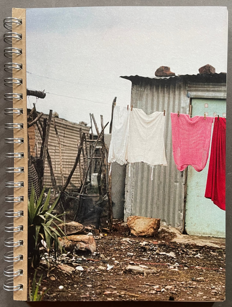 A spiral-bound notebook with a photographic cover depicting a rustic outdoor scene with laundry hanging on a line, set against a dark gray background.