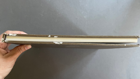 A hand holding the side edge of a thick, vintage-style book or album with a brown cover and cream-colored pages, set against a plain gray background.