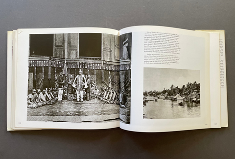 An open vintage book laid flat on a dark gray surface, showing two printed pages with black-and-white historical photographs and text. The left page contains a large scene with a group of people gathered indoors, and the right page contains a smaller landscape photograph with water and boats.