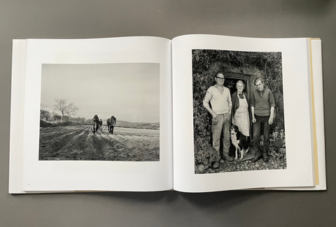 An open hardcover book laid flat on a dark gray surface, showing two black-and-white photographic prints on the pages. The left page contains a landscape photo of two people riding horses along a dirt road, and the right page shows a black-and-white portrait of three people standing outdoors with a dog.
