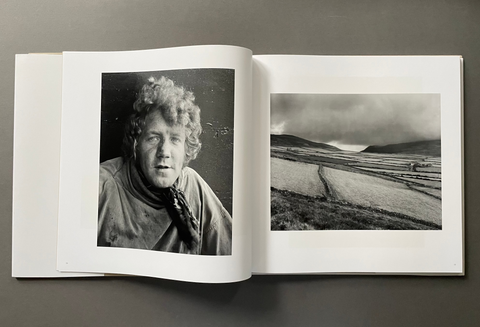 An open hardcover book or photo journal laid flat on a dark gray surface, showing two black-and-white printed photographs on white pages: a portrait on the left page and a landscape scene on the right page.