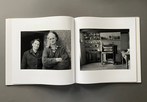 An open hardcover book laid flat on a dark gray surface, showing two black-and-white photographs one with a woman and man posing and house interior printed on the pages.