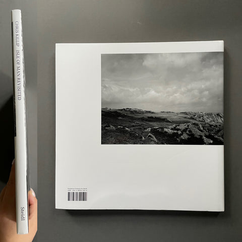 A hardcover art book titled 'Isle of Man Revisited' by Chris Killip, held by a hand to show its spine alongside the white front cover featuring a black and white landscape photograph. The book is set against a dark gray background.