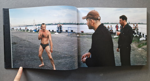 An open hardcover book, showing a large photographic scene: a shirtless man in swim trunks on the left and two men standing by the water looking at him on the right.