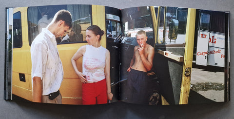 An open book spread, showing a man in a white shirt and a woman in a white top and red pants standing beside a yellow vehicle. There is another man next to them smoking a cigarette.