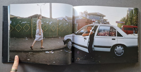 An open book showing a printed photograph of a woman walking beside a white car on a street, held open by a hand of a man. Car is full with apples.