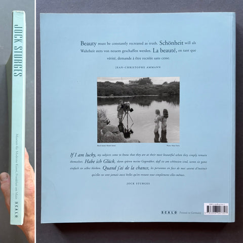 The back cover of the book 'Beauty Must Be Constantly Recreated' by Jock Stürmers, held by a hand on the left side to show the book's thickness. The cover is a pale blue with German text, a central black-and-white photograph of two people by a lake, and a light blue spine visible on the left.