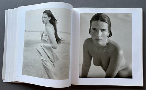 An open book or photo journal laid flat on a dark surface, showing two black-and-white photographic prints side by side. The left page features a beach scene with a woman wrapped in a towel, and the right page shows a close-up portrait of a nude woman.