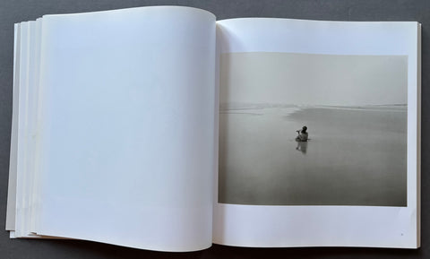 An open book or photo journal laid flat on a dark gray surface, showing a large white left page and an open right page with a minimalist black-and-white landscape photograph of a small figure in a misty flat landscape.