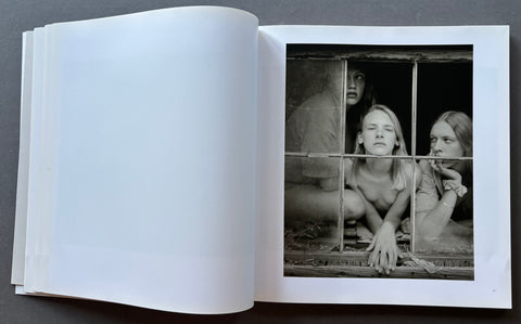 An open art book or photo journal laid flat on a dark surface, showing a large black-and-white photographic print on the right page with three seated figures behind a barred window, and an empty white left page.