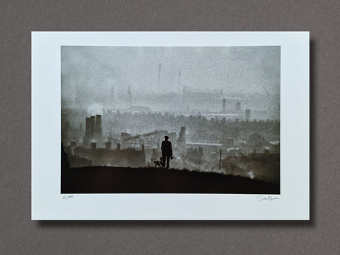 A framed or matted art print showing a misty industrial cityscape with a small dark silhouette of a person standing on a hill overlooking the skyline. The print is photographed against a plain taupe-gray background.