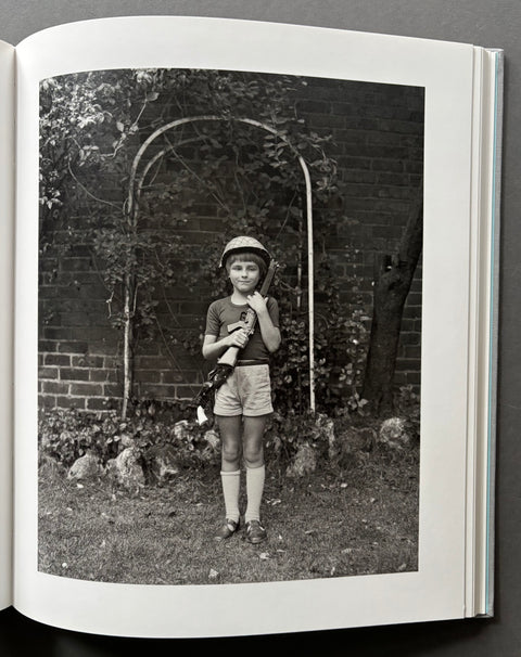 An open book or photo album showing a black-and-white printed photograph on the page. The photo depicts a child standing outdoors in front of a brick wall and arched trellis, holding a camera.