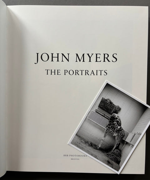 The front cover of the book 'John Myers: The Portraits' by Red Photographic Studio, featuring a minimalist white design with black text and a tilted black-and-white portrait of a giraffe.