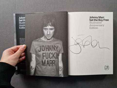 An open book showing the cover and interior pages of 'Johnny Marr: Set the Boy Free Illustrated Anniversary Edition', held by a hand against a dark gray background. The left page features a black-and-white portrait of Johnny Marr wearing a t-shirt with the text 'JOHNNY FUCKIN MARR', and the right page shows a printed title and a large autograph.