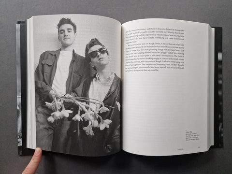 An open hardcover book held by a hand, showing a black-and-white photograph of two men on the left page and printed text on the right page, against a dark gray background.