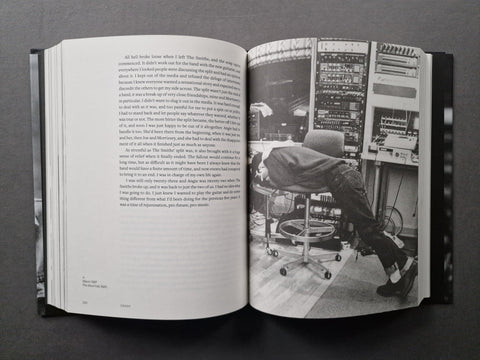 An open hardcover book laid flat on a dark surface, showing a two-page spread with printed text on the left page and a black-and-white photograph on the right page depicting a person seated in front of a control panel.