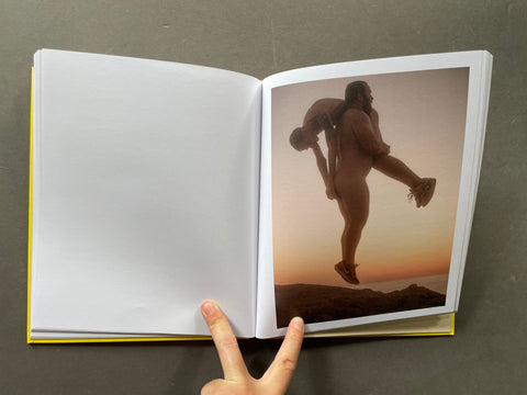 An open hardcover book with blank white pages on the left and a printed photograph on the right showing two people outdoors at sunset. A hand is visible holding the book open against a dark gray background.