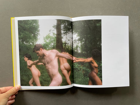 An open art book held by a hand, displaying a photographic print of several nude figures in a forest on the left page and a blank white page on the right, set against a dark gray background.