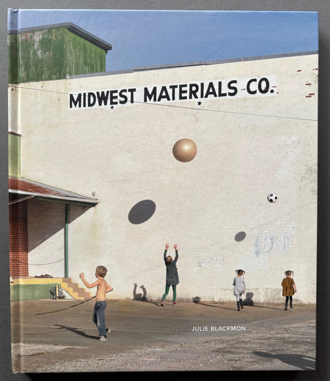 Book titled 'Midwest Materials' by Julie Blackmon with a cover featuring the image of children playing in front of a building with the same name as the title of the book.