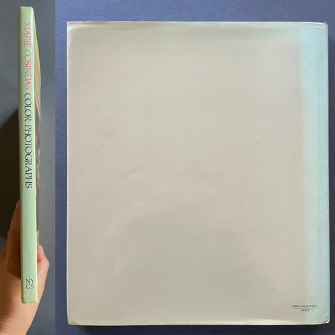 A hand holds the spine of a book titled 'Marie Cosindas COLOR PHOTOGRAPHS' next to the blank back cover of the same book, set against a dark blue background.