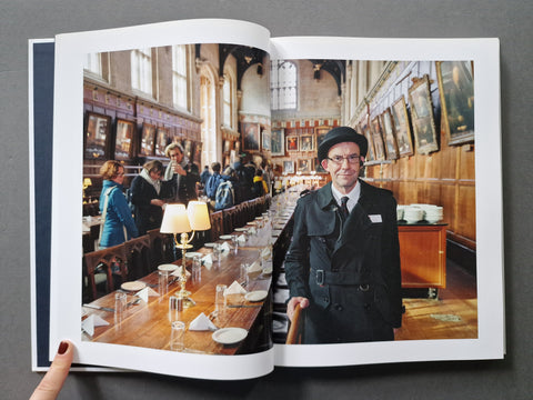 An open hardcover, showing a large printed photograph of a man in a black coat and hat standing in a long dining hall with people seated along tables.