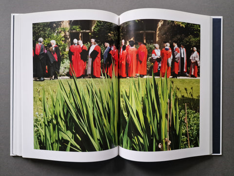 An open hardcover book, displaying a double-page spread with a large photographic print. The print features a group of people in red and black academic robes standing outdoors on a grassy lawn, with tall green reeds in the foreground.