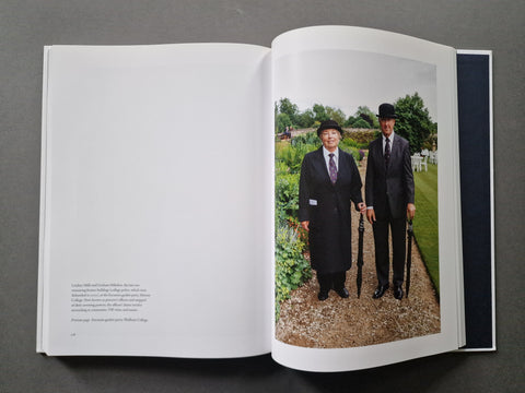 An open book, showing a white left page with small text and a large right page photograph of two men in black suits standing outdoors on a gravel path with greenery behind them.