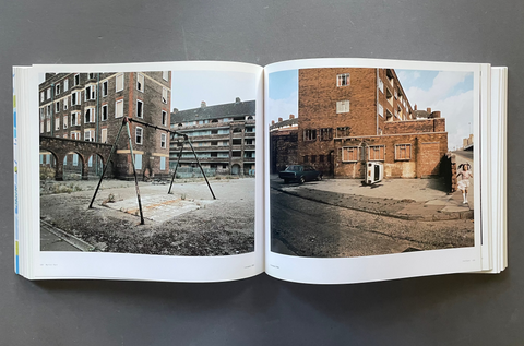 An open book laid flat on a dark gray surface, showing two printed photographs side by side. The left page features a black-and-white photo of an urban courtyard with a swing set and brick buildings, while the right page shows a color photo of a brick building courtyard with a car and a small figure.