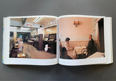 An open book or photo album laid flat on a dark gray surface, showing two interior photographs printed on the pages. The left page depicts people sitting at slot machines in a casino-like room, and the right page shows a couple seated at a table in a restaurant or cafe with pink walls.