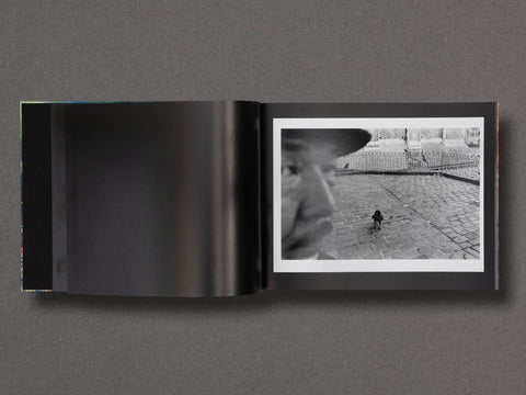 An open black-and-white photo book or art print set displayed flat against a dark gray background. The left page is mostly dark, while the right page shows a black-and-white photograph of a person in profile and a small bird on a cobblestone surface.