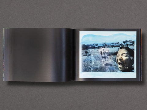 An open hardcover book laid flat on a dark gray surface, showing a printed interior spread with a large black left page and a blue-toned photographic page on the right featuring a screaming face and outdoor landscape.