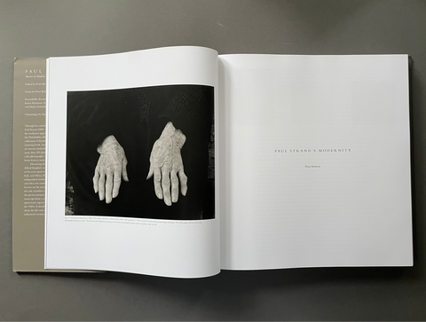 An open book or art print laid flat on a dark gray surface, showing a black-and-white photograph of two pale hands on the left page and small centered text on the right page.