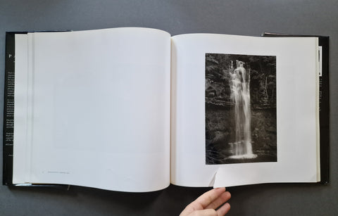 An open hardcover book or photo album laid flat on a dark gray surface, showing a black-and-white waterfall photograph printed on the right page. A hand is visible holding the bottom edge of the page.