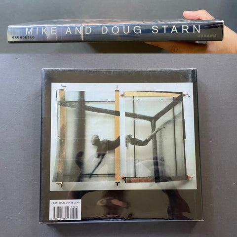 A hardcover book titled 'Dreams' by Mike and Doug Starn, shown with the top cover held by a hand above the closed bottom cover, on a dark gray surface.