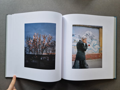 An open book showing two printed photographs on the inner pages: one of a flowering tree at dusk and one of a woman standing in front of a graffiti wall holding a red flower.