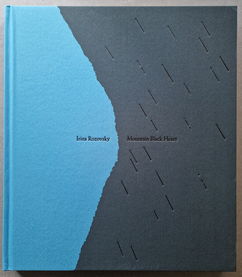 A square hardcover book titled 'Mountain Black Heart' by Irina Rozovsky, featuring a minimalist cover design with a vertical blue-to-black gradient and small cutout rain-like lines on the black side.