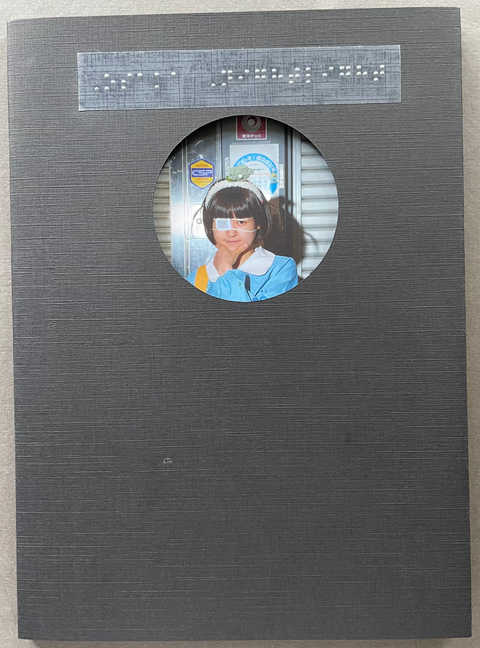 A square dark gray textured book or album cover lies on a light beige surface. The cover features a circular cutout in the center showing a young girl with dark hair wearing a light blue jacket, and a strip of white Braille text near the top.