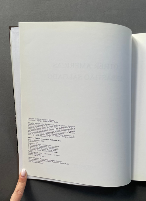 An open book or binder showing a blank white page with faint embossed text and a block of small printed text below, held open by a hand against a dark gray background.