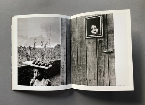 An open book or photo journal laid flat on a dark gray surface, showing two black-and-white photographic prints on the spread. The left page depicts a woman carrying fruit on her head outdoors, and the right page shows a framed portrait of a woman on a weathered wooden wall.