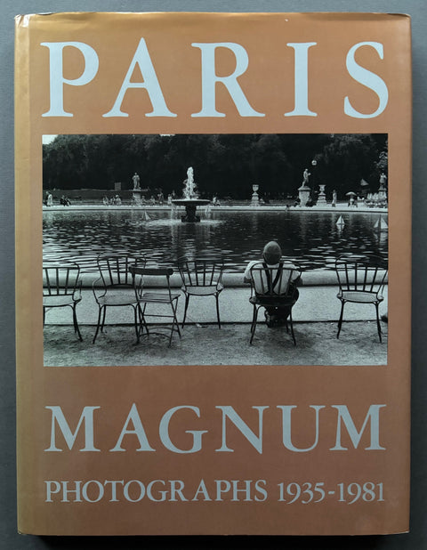 Book titled 'Paris: Magnum Photographs 1935-1981' with a beige cover featuring an image of a man sitting on a chair next to a pond with a fountain.