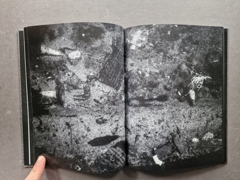 An open book showing a black and white photographic print of debris or rubble, photographed against a gray background with a finger pointing at the bottom left.