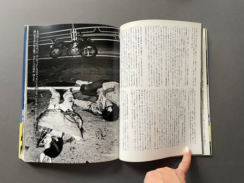 An open Japanese book or magazine lying on a dark gray surface, showing a black-and-white photograph of people on the left page and dense text on the right page. A person's hand is holding the open book at the bottom right.