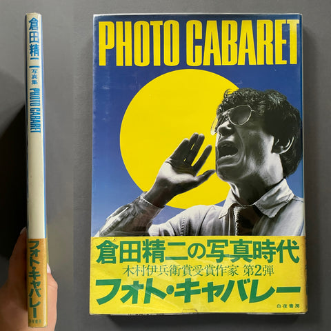 A hardcover book titled 'Photo Cabaret' shown standing upright next to its spine, with a hand holding the spine for scale. The cover features a large yellow sun graphic, bold yellow title text, and a black-and-white portrait of a man wearing glasses against a blue background.
