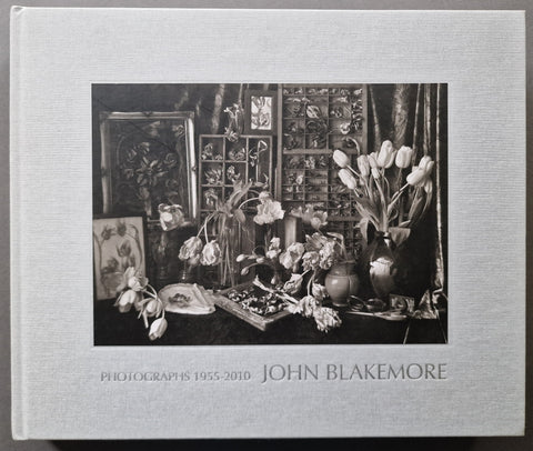 A hardcover art book titled 'Photographs 1955-2010 John Blakemore' featuring a black and white still life photograph of flowers and framed art on the cover, set against a light gray background.