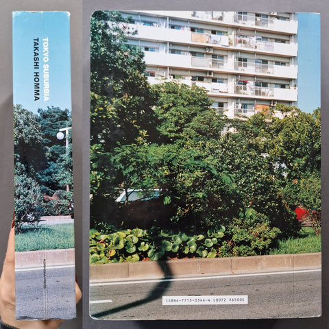 A hardcover book titled 'Tokyo Suburbia' by Takashi Homma, held by a hand. The cover features a vintage photograph of a Tokyo apartment building surrounded by lush green trees and a road.