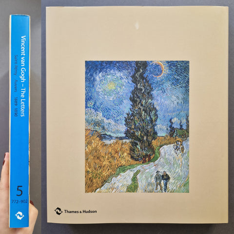 A hardcover book titled 'Vincent van Gogh - The Letters' by Thames & Hudson, shown standing upright next to a hand holding its blue spine. The front cover features a central reproduction of Van Gogh's 'Starry Night' painting on a beige background.