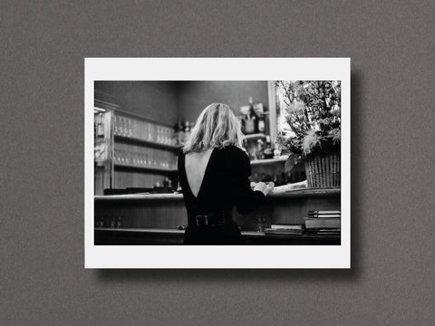 A black and white photographic art print featuring a woman seen from behind at a bar, displayed with a wide white border against a textured gray background.