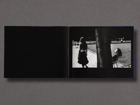 An open black book laid flat on a gray surface, showing a black-and-white photograph printed on the right page with a silhouetted figure walking beside a tree in an outdoor park setting.